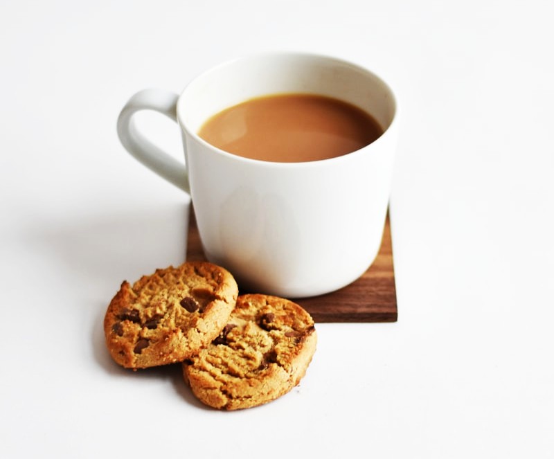 White mug of coffee with chocolate chip cookies on a wooden coaster